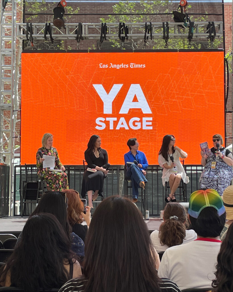 four women on a stage. They are all holding books; Amy Spalding (on the right of the photo) is holding up her book, _No Boy Summer_. A huge orange screen with "YA Stage" is lit up behind them all. 